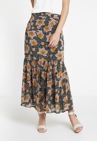 Schwarz gemusterter Midirock mit floralen Motiven in Pink und Orange, kombiniert mit weißen Sandalen mit Knöchelriemen und einem weißen ärmellosen Top.