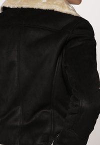 Svart fårskinnsjacka med en krämfärgad pälskrage, strukturerad tyg, figursytt snitt och synliga sömdetaljer på ryggen och ärmarna.
