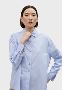 Camicia azzurra chiara con righe verticali bianche, caratterizzata da scritte ricamate e un colletto classico, realizzata in tessuto morbido.