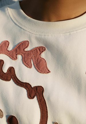 Valkoinen collegepaita, jossa on monimutkaisia kangasapplikaatioita vaaleanpunaisen ja ruskean sävyissä, resorilla viimeistelty pyöreä pääntie ja erottuvat ompelut.