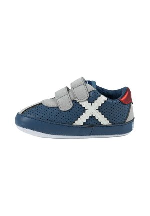 Zapatilla infantil perforada azul marino con tiras de velcro grises, detalle en forma de X blanco cosido y acento rojo en el talón, vista lateral.