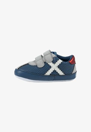 Zapatilla infantil perforada azul marino con tiras de velcro grises, detalle en forma de X blanco cosido y acento rojo en el talón, vista lateral.