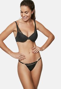 Soutien-gorge en dentelle noire avec des bonnets rembourrés et des bretelles décoratives, associé à un string en dentelle noire assorti avec une bande accentuée et des bretelles latérales fines.
