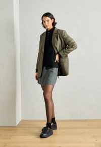 Blazer verde oliva sopra un maglione nero a trama, abbinato a una gonna verde, collant neri trasparenti e sneakers nere con suola spessa.