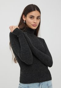 Maglione a collo alto ribassato di colore grigio scuro, con maniche lunghe. Presenta un design aderente e un tessuto testurizzato. Indossato con jeans in denim azzurri.