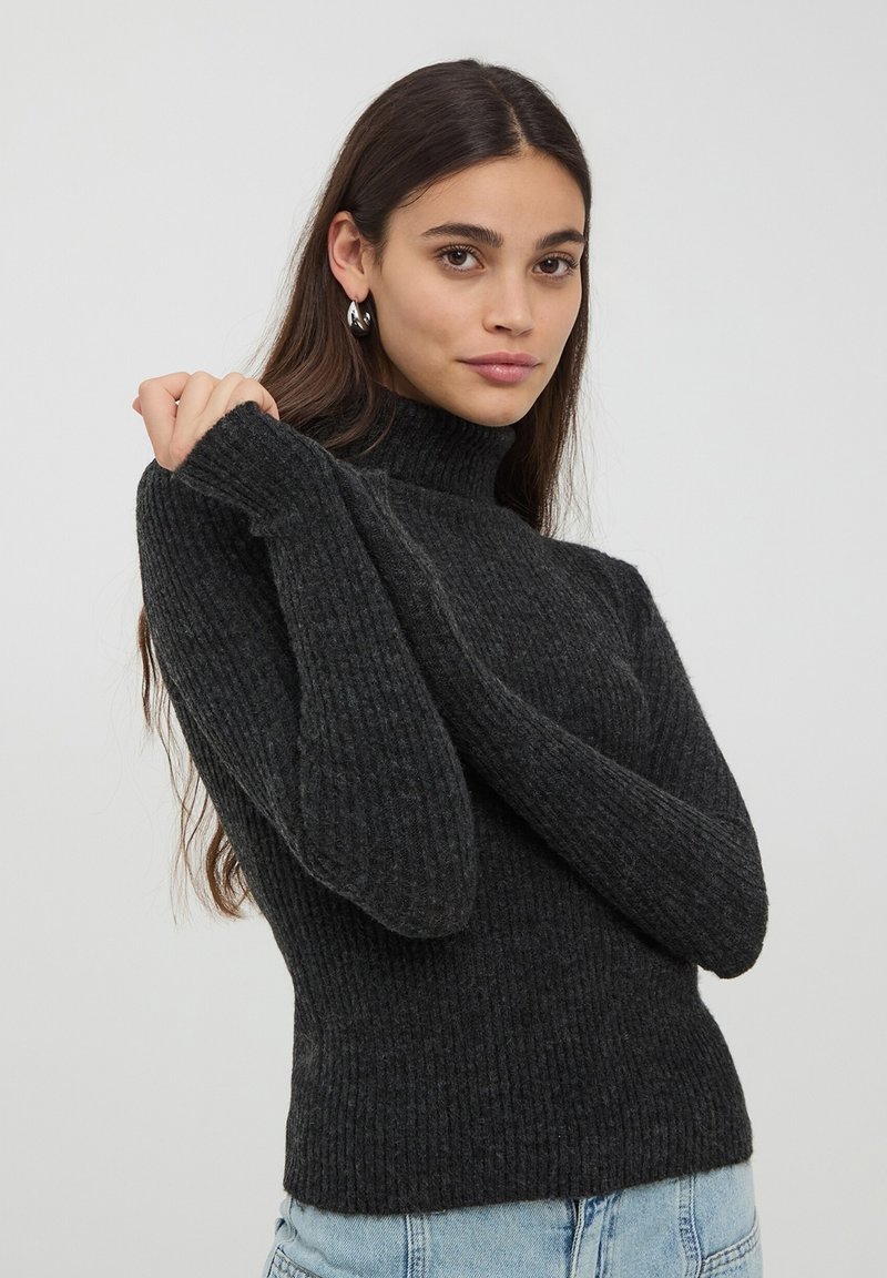 Maglione a collo alto ribassato di colore grigio scuro, con maniche lunghe. Presenta un design aderente e un tessuto testurizzato. Indossato con jeans in denim azzurri.