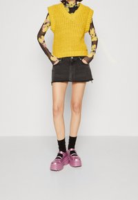Geel gebreid mouwloos topje met V-hals, gecombineerd met een zwarte denim minirok. Afgemaakt met roze platform schoenen en zwarte enkelsokken.