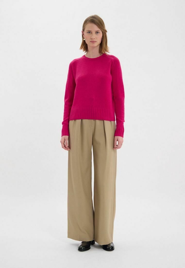 Gilet en tricot fuchsia avec un col rond et des poignets côtelés, associé à un pantalon large beige et des chaussures noires. Lignes simples et épurées.