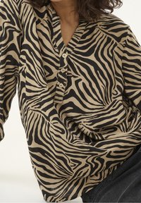 Chemise beige à boutons avec des rayures zébrées noires, dotée d'un col en V et d'un tissu doux et léger. Les détails comprennent de petits boutons et une coupe décontractée.