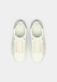 Baskets basses blanches avec motif texturé, lacets beiges et accents argentés au talon, vues de dessus sur un fond uni.