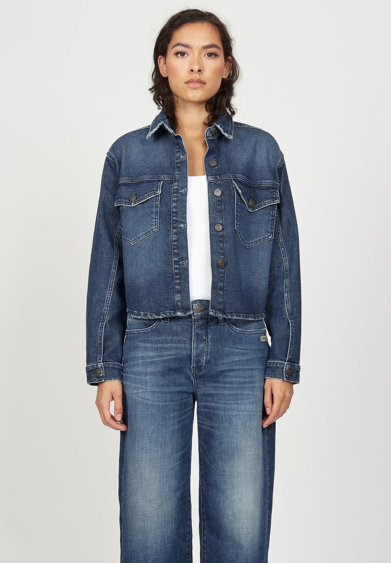 Jeansjacke in Dunkelblau, mit knöpfbarem Vorderteil, zwei Brusttaschen und kontrastierenden Nähten. Getragen über einem weißen Oberteil und kombiniert mit blauen Jeans.