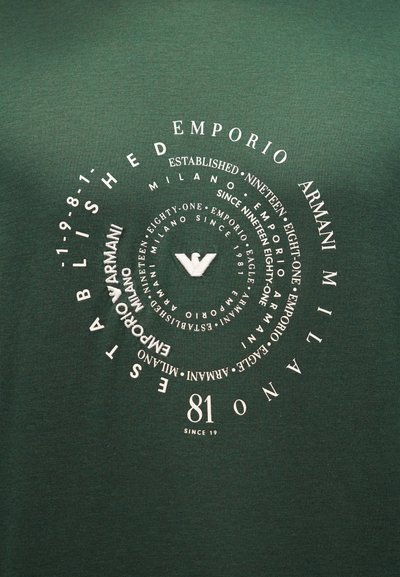 Πράσινο ύφασμα με λευκό κείμενο διατεταγμένο σε συγκεντρικούς κύκλους, που περιλαμβάνει τα «Emporio Armani Milano», «Established 1981» και ένα κεντρικό λογότυπο αετού.