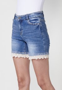 Gaišas zila džinsu šorti ar baltām šuvēm. Iekļauj viļņotu mežģīņu apakšmali un divas priekšējās kabatas. Klasiska piecu kabatu dizains.