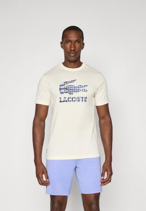 T-shirt crème avec un imprimé graphique bleu d'un crocodile et le texte "LACOSTE". Manches courtes et col rond. Shorts bleu clair.
