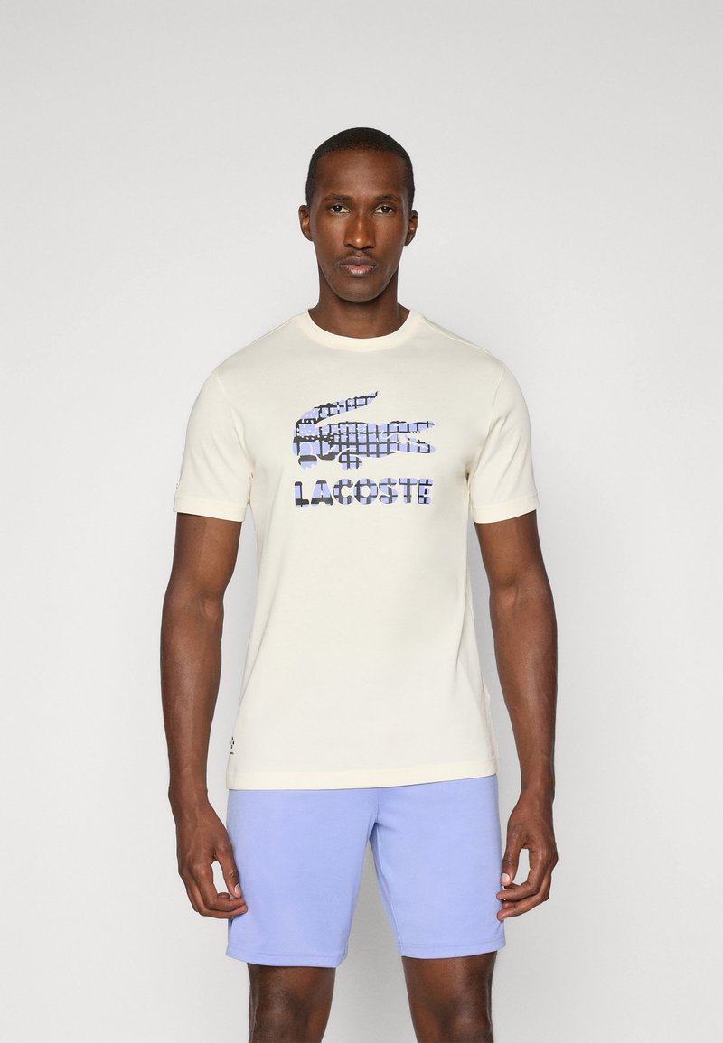 Krém színű póló, amelyen kék grafikai nyomat látható egy krokodilról és a "LACOSTE" feliratról. Rövid ujjú és kerek nyakú. Világoskék rövidnadrág.