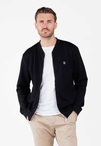 Hombre con barba clara y bigote, vestido con chaqueta negra con cremallera sobre una camiseta blanca, pantalones beige, de pie con las manos en los bolsillos.