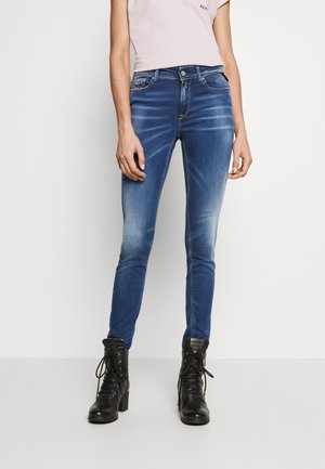 Kobieta w wyblakłych niebieskich obcisłych dżinsach z przednimi kieszeniami i czarnych sznurowanych botkach, stojąca na białym tle.