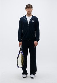 Sudadera con capucha azul marino con cremallera y detalles blancos, pantalones a juego y zapatillas blancas. Sosteniendo una raqueta de tenis con detalles en amarillo y púrpura.