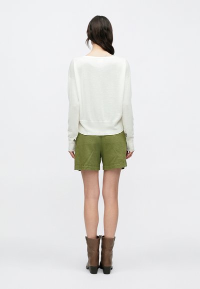 Γυναίκα που στέκεται με την πλάτη προς τα έξω, φορώντας λευκό μακρυμάνικο τοπ, χακί σορτς και καφέ μποτάκια μέχρι τη μέση της γάμπας, σε απλό φόντο.