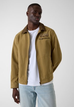 Jeune homme portant une veste beige zippée avec la broderie « GUESS Country Club », un t-shirt blanc et un jean bleu clair, debout devant un fond gris.