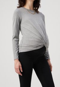 Mujer embarazada que lleva una blusa gris de manga larga con un detalle de nudo en la parte delantera y pantalones de maternidad negros, de pie frente a un fondo sencillo.