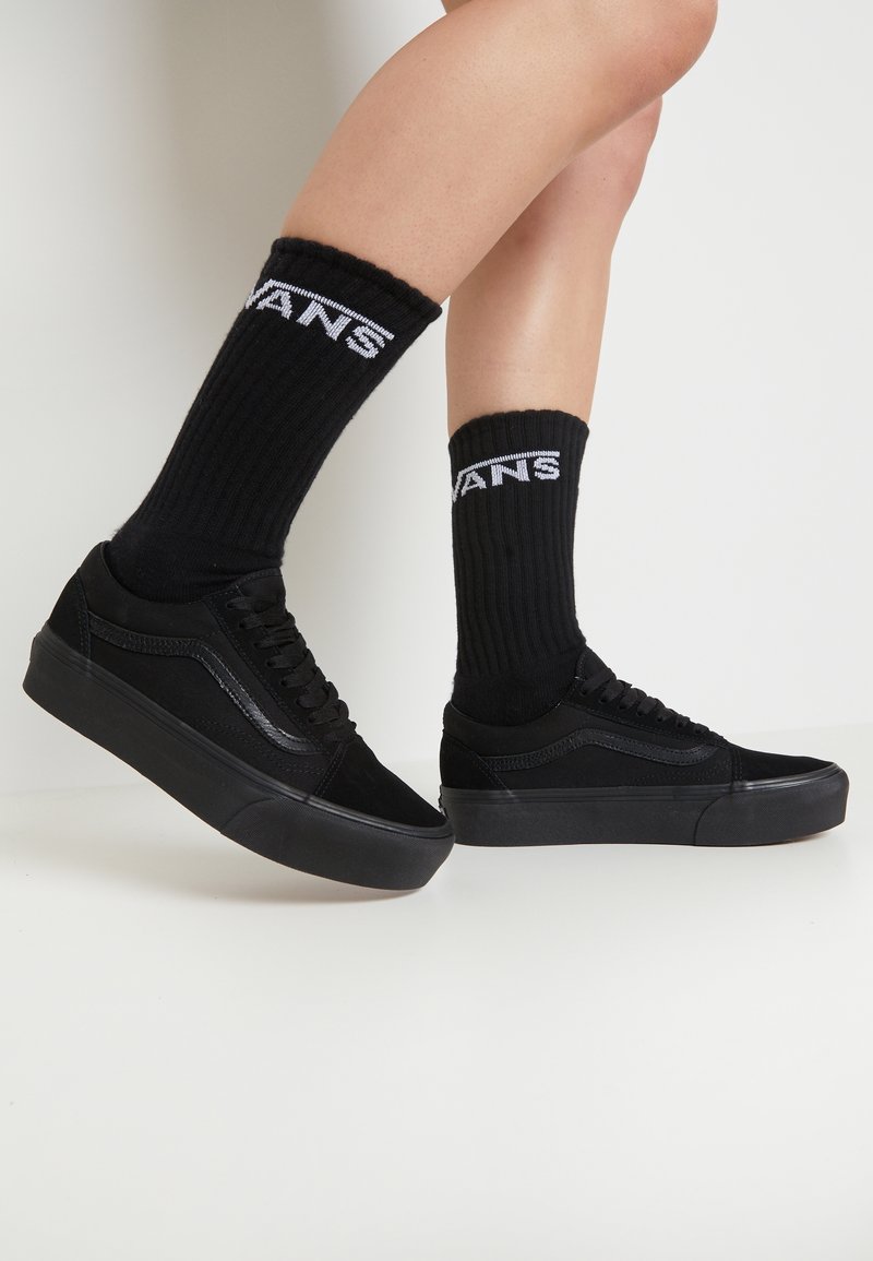 Jambes portant des baskets Vans noires et des chaussettes côtelées noires avec le logo Vans blanc sur fond blanc.