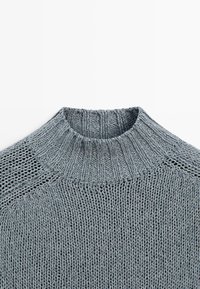 Grå strikket sweater med ribbet halsrør og blandede strikkemønstre på øverste del af brystet og ærmerne.