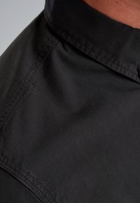 Camicia nera con colletto, realizzata in tessuto liscio e opaco. Presenta cuciture visibili e un design dal taglio rilassato. Vista ravvicinata del colletto e della texture del materiale.