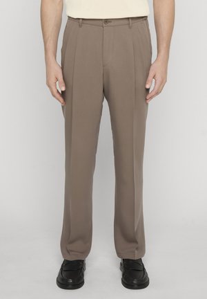 Homme portant un pantalon plissé beige et des mocassins noirs, debout devant un fond uni.