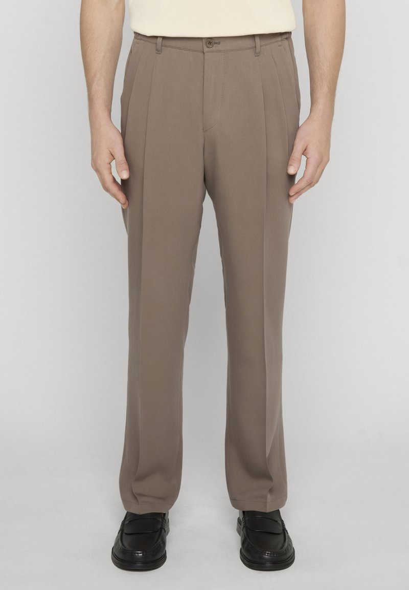 Homme portant un pantalon plissé beige et des mocassins noirs, debout devant un fond uni.