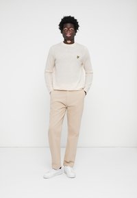 Pull beige à col rond avec un petit logo jaune sur la poitrine, associé à un pantalon beige clair et des baskets blanches.