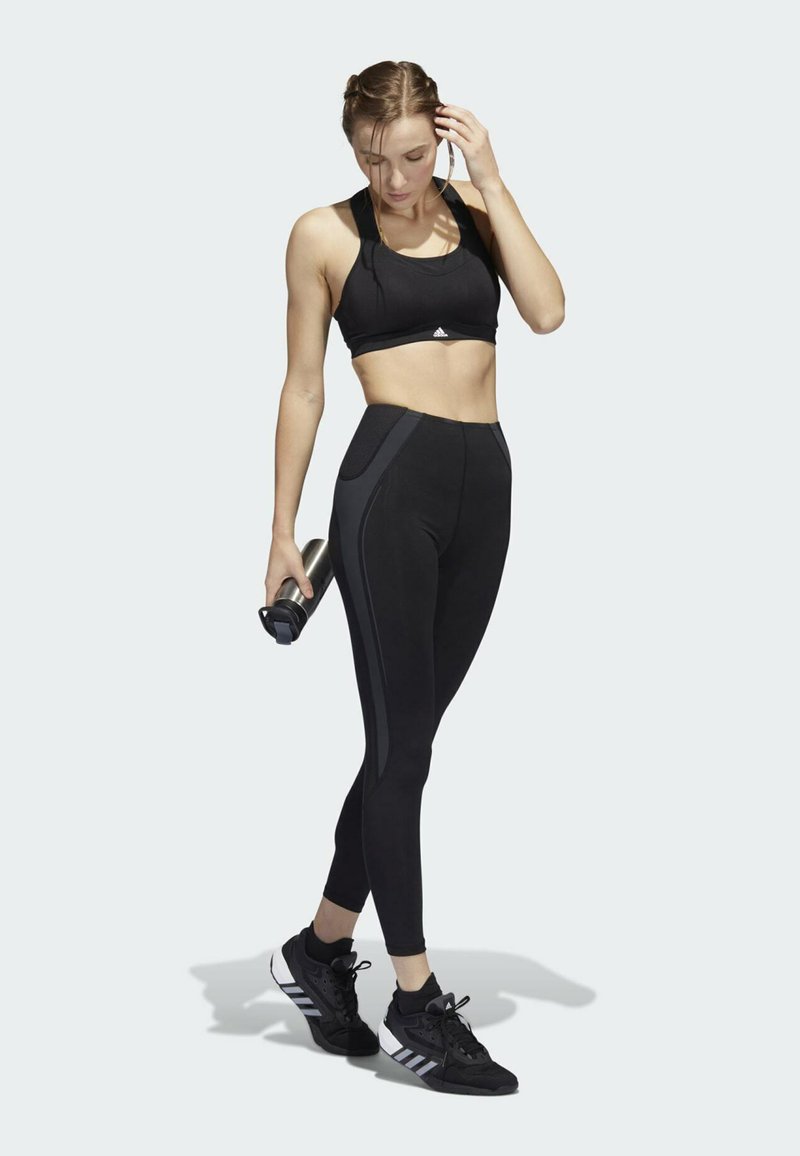 Schwarzer Sport-BH und hoch geschnittene Leggings, beide figurbetont, mit kontrastierenden Seitenpaneelen. Schwarze Sportschuhe mit weißen Akzenten. Hält eine Wasserflasche.