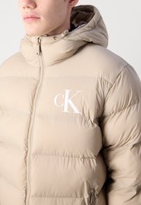 Beige puffer jacket with a hood, featuring a front zipper and a white logo on the chest. Smooth texture, quilted design, and elastic cuffs.