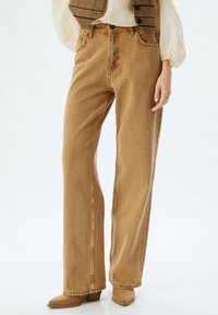 Jeans de pierna recta de color marrón claro, hechos de mezclilla, con bolsillos frontales, passantes para cinturón y una textura ligeramente deslavada.