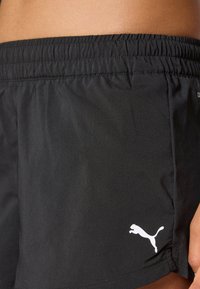 Närbild av en person som bär svarta Puma-idrottsshorts med elastisk midja och Puma-logotyp på vänster lår.