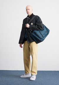 Palton negru cu multiple buzunare, cămașă verde, pantaloni bej și adidași albi. Poartă o geantă albastră cu clapetă și detalii cu logo.