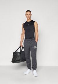Sort ærmeløs Nike top, grå sweatpants med hvidt logo og hvide atletiske sko. Holder en sort træningstaske. Enkel, sporty design.