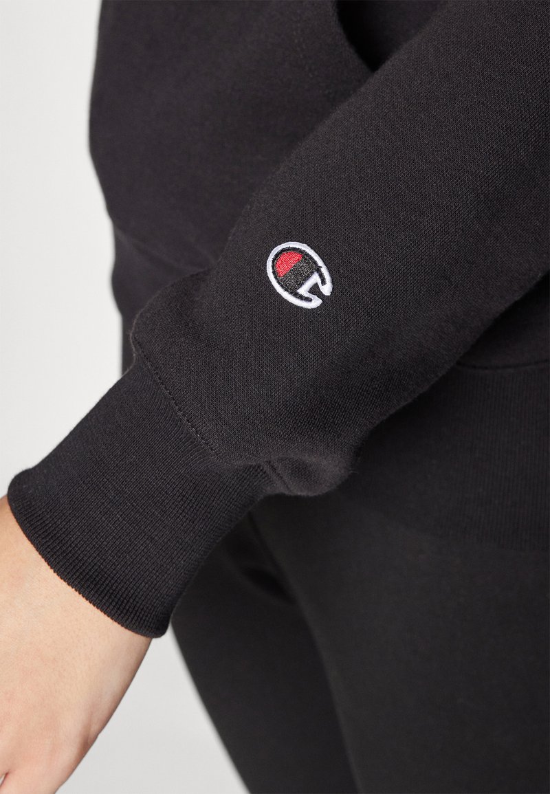Schwarzer Sweatshirt mit gerippten Bündchen, ausgestattet mit einem kleinen gestickten Logo in Rot und Weiß auf dem linken Ärmel. Weiche Stofftextur.