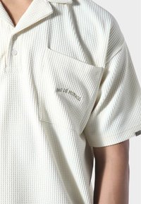 Polo tričko s krátkým rukávem z texturované, krémově bílé látky; obsahuje límec, přední kapsu a vyšité "PAS DE MONACO" v hnědé barvě.