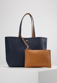 Sac fourre-tout bleu marine avec de longues poignées et un petit logo en argent, associé à une pochette en cuir brun texturé détachable placée devant.