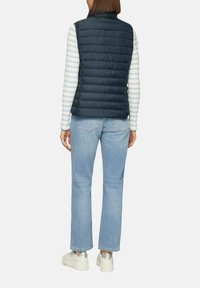 Gilet matelassé marine, porté sur un haut à manches longues rayé, associé à un jean bleu clair et des baskets blanches imposantes.