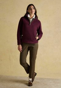 Burgundijas siltā vilnas džemperis ar pusvīles apkakli, sānu kabatām, kopā ar brūniem rūtainiem biksēm un brūnām kurpēm, uz neitrāla fona.