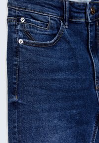Denim jeans featuring a dark blue wash, five-pocket design, and contrasting stitching. Includes metal rivets on pocket edges.