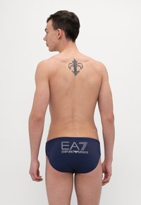 Tengerészkék úszóalsó, kontrasztos ezüst EA7 és Emporio Armani logókkal a hátsó részen. Sima anyag, testhezálló fazon, minta vagy díszítés nélkül.