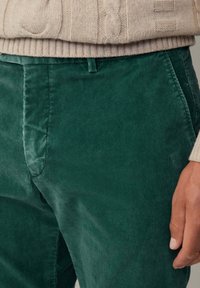 Pantalons en velours côtelé vert avec des côtes verticales, une texture lisse et une coupe ajustée avec un détail subtil à la taille. Associés à un pull en tricot beige.
