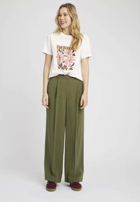 T-shirt blanc avec imprimé floral en rose et marron, associé à un pantalon large vert olive. Le modèle porte des baskets bordeaux.