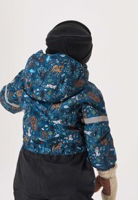 Sininen kuviollinen toppatakki, jossa eläimiä ja puita, musta hattu ja lapaset. Hihoissa huomiota herättävä heijastin.