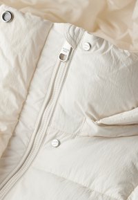 Chaqueta acolchada color beige claro con un acabado suave y texturizado. Cuenta con un cierre central de cremallera, botones de presión y detalles de hardware de marca.