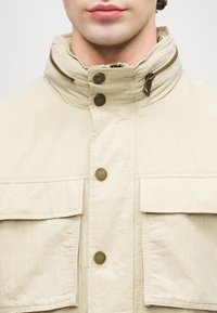 Vyras su béžiniu švarku, turinčiu aukštą apykaklę, dvi priekinės kišenes su klostėmis, metaliniais paspaudimo mygtukais ir užtrauktuku šalia apykaklės.