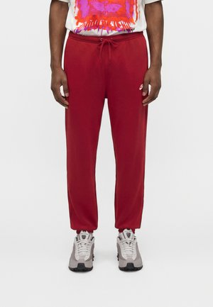 Person iført røde Nike træningsbukser med snøre, hvide og grå Nike sneakers og en hvid T-shirt med et abstrakt rødt og lilla grafisk motiv.